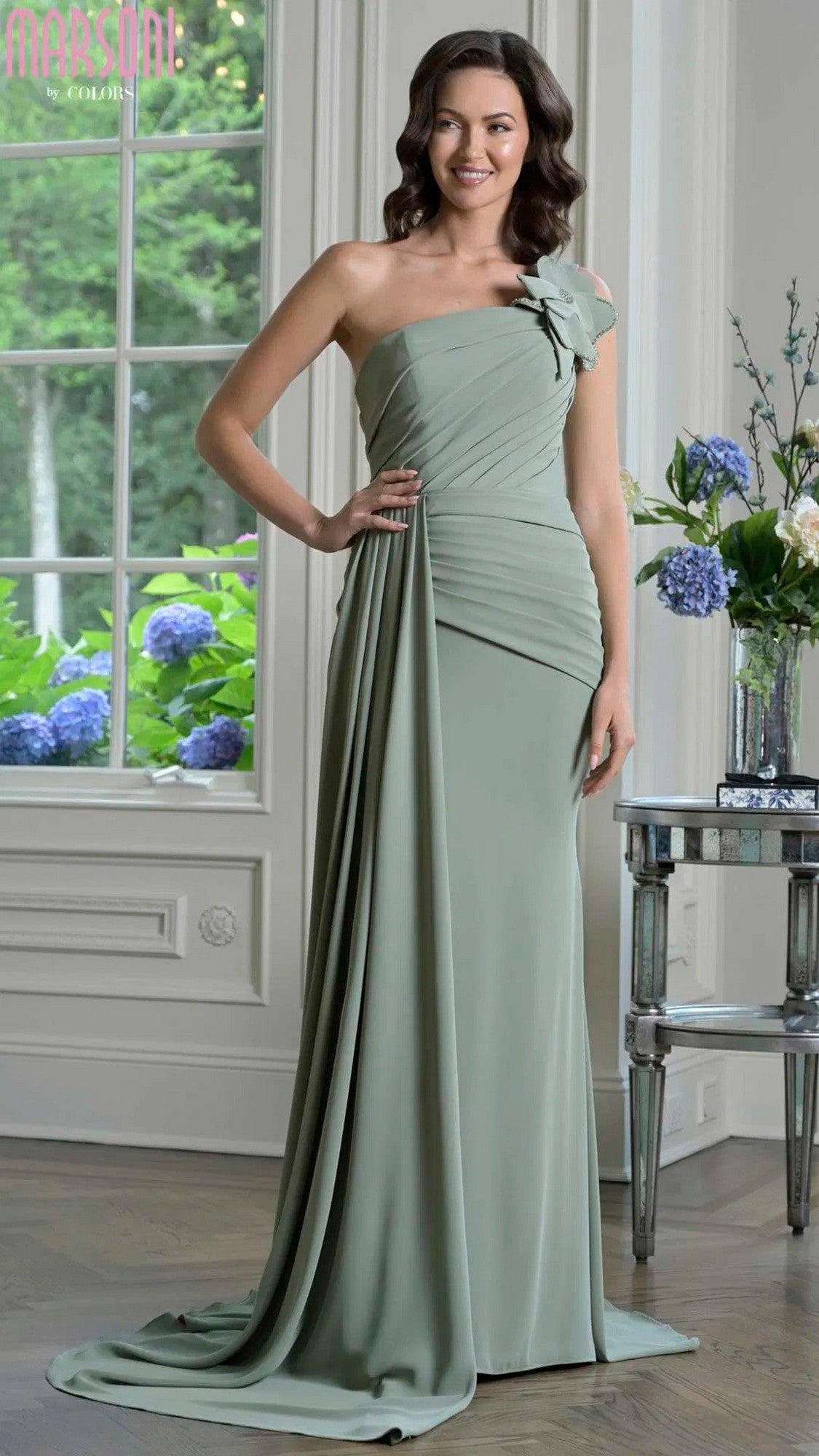 A woman wearing the Marsoni M330 long formal dress in sage, featuring a one-shoulder design and draped fabric, stands indoors near windows beside a table with floral arrangements.